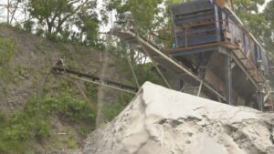 Tampilan wide shot mesin stone crusher dan conveyor belt yang sedang mengalirkan material abu batu ke dalam gundukan besar